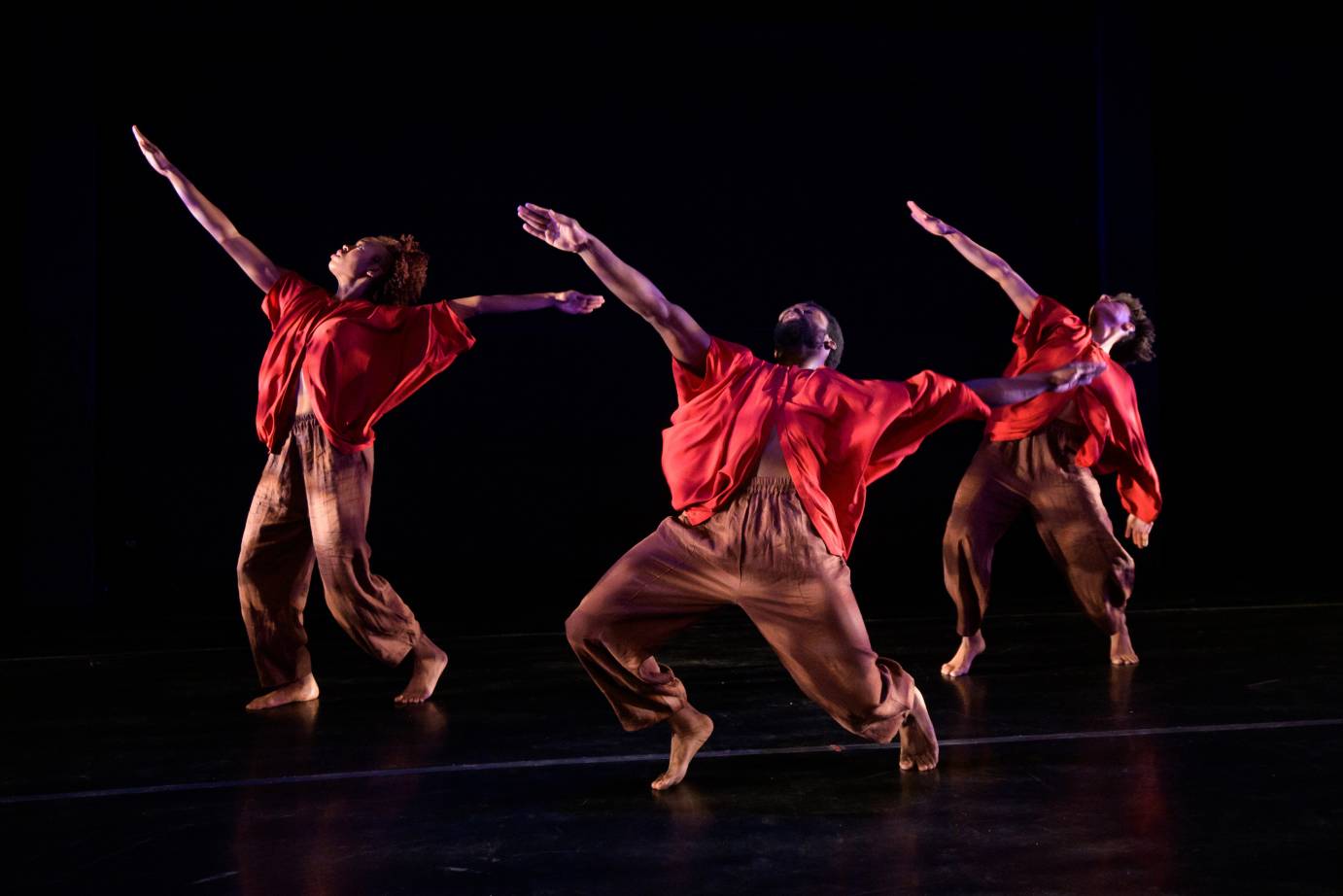 Three dancers in red tops extend an arm on the diagonal, their knees bent